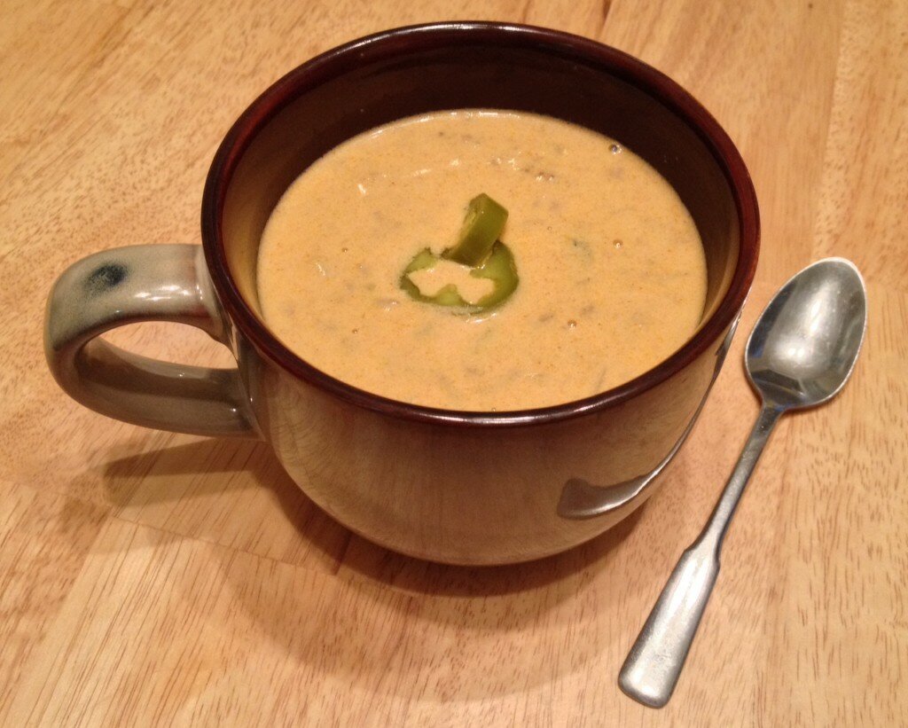 Homepage post card 3: Creamy orange beef nacho soup in a dark brown mug garnished with a jalapeño ring, spoon beside it on wood