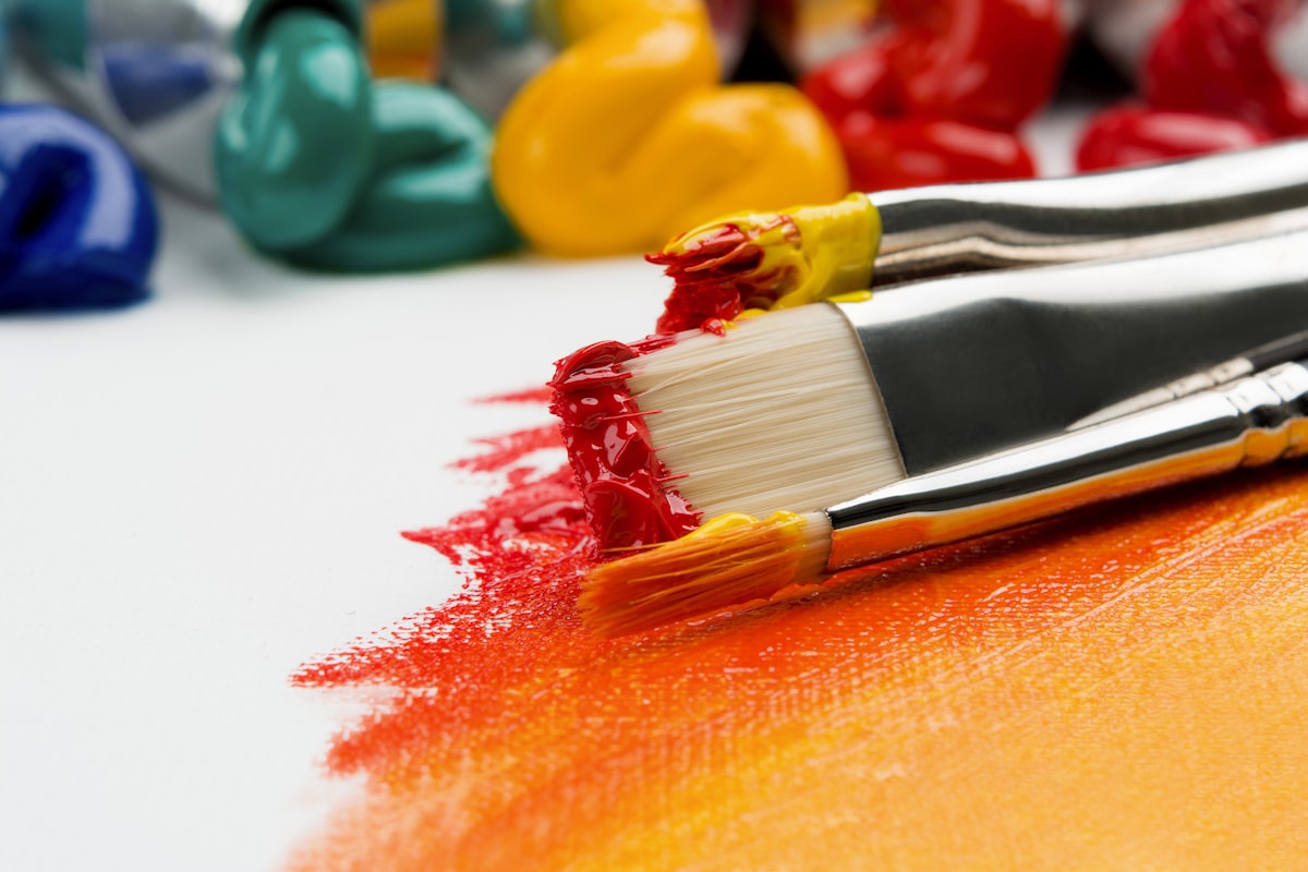 Pagination page 3 card 1: Close-up of paintbrushes loaded with wet red and yellow paint beside blue, teal, and yellow paint swirls on white